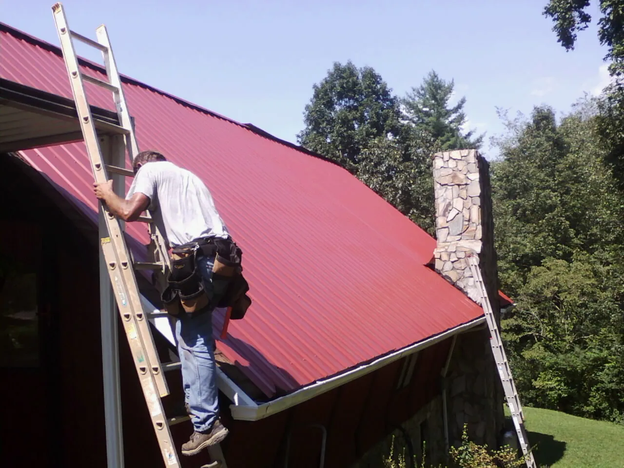 Roofer on a ladder beside a red metal roof and stone chimney — Soffit & Fascia Repair by Roofing Brothers in Gifford